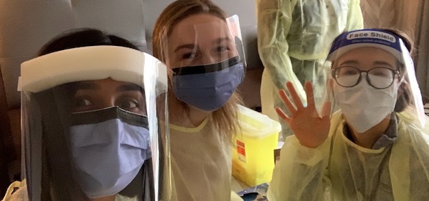 Three female doctors wearing personal protective equipment wave to the camera.