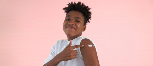A young person has their sleeve rolled up pointing at a bandaid on their arm where they received a vaccination.